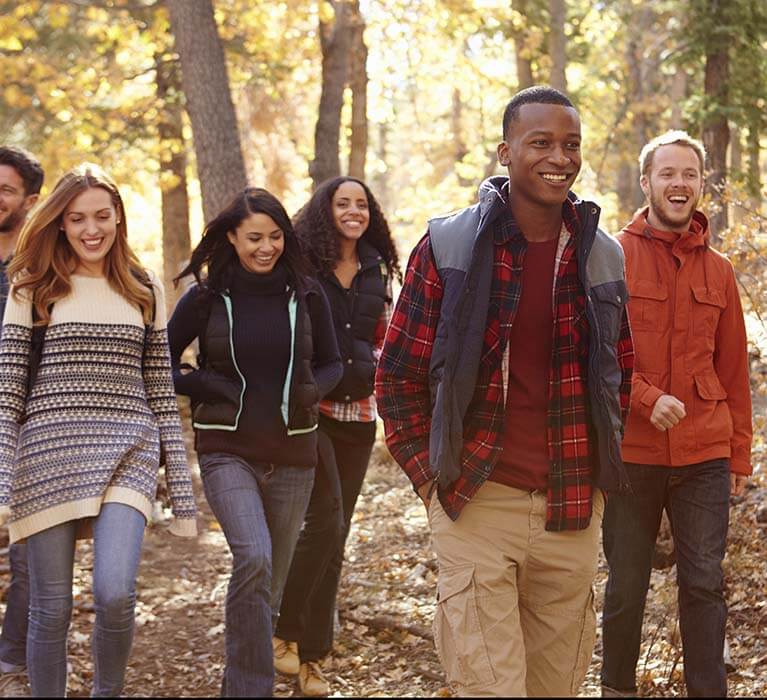 A group of young adults, dressed in casual fall clothing, smile and walk together on a wooded trail surrounded by autumn foliage.