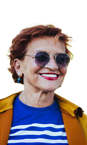 An older woman with short, curly red hair smiles brightly. Confident despite her hearing loss, she wears round sunglasses, a blue and white striped shirt, and a mustard yellow jacket, standing against a plain white background.