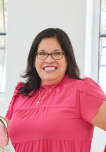 A woman with dark hair and glasses, wearing a bright pink blouse and a necklace with a cross pendant, smiles at the camera in a brightly lit room.