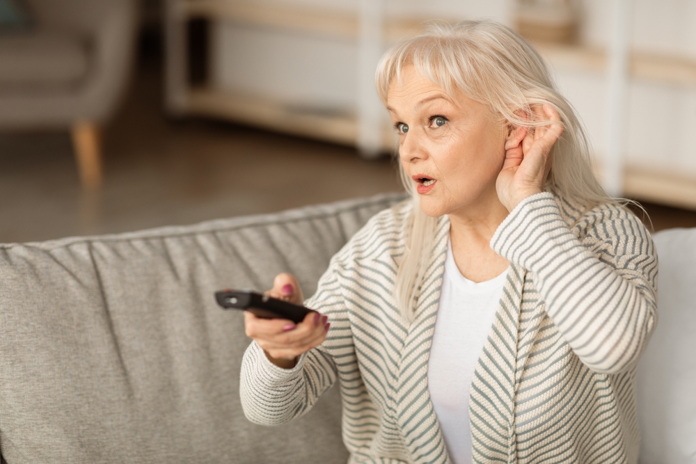 An older woman sitting on a sofa holds a TV remote in one hand and cups her ear with the other, appearing to have difficulty hearing. She looks focused and is wearing a striped cardigan.
