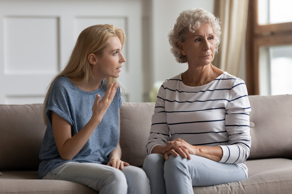 A younger woman sits on a couch speaking emotionally to an older woman with gray hair, who looks away with a serious, distant expression. Both women appear tense and upset.
