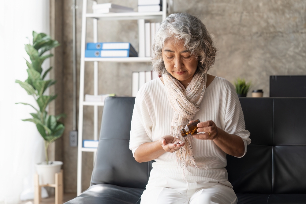 An older woman with gray hair sits on a black sofa, pouring pills from a brown bottle into her hand. She wears a white outfit and a scarf, with a bookshelf and plant in the background.