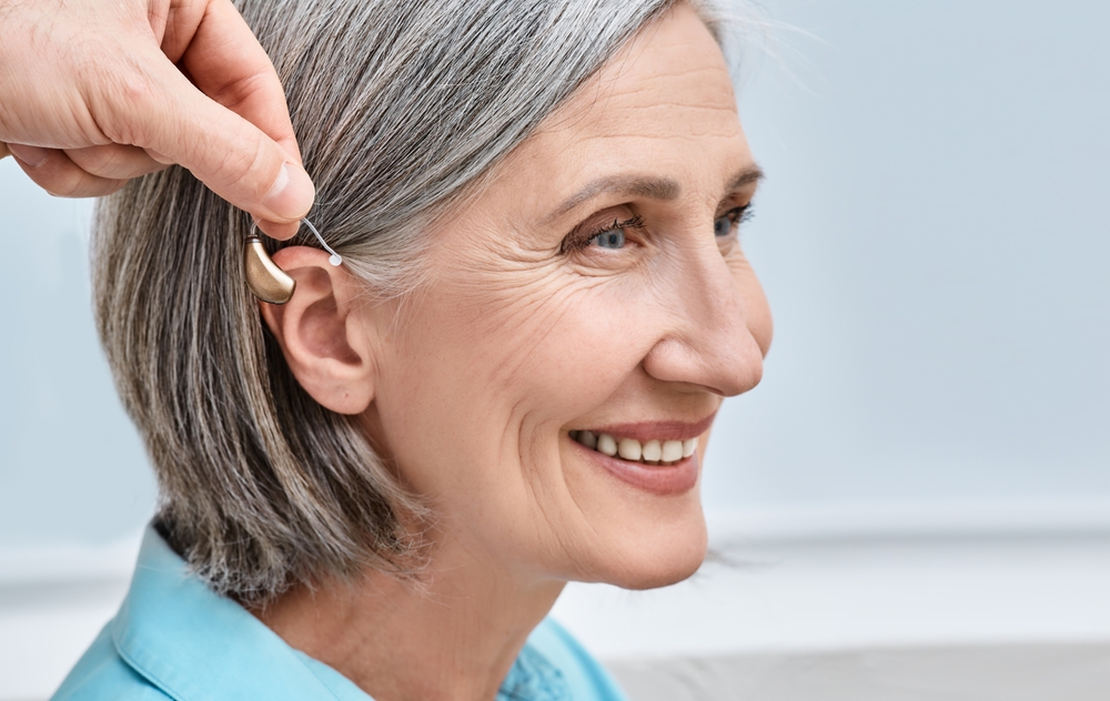 A smiling older woman with gray hair has a hearing aid being placed behind her ear by another person’s hand.