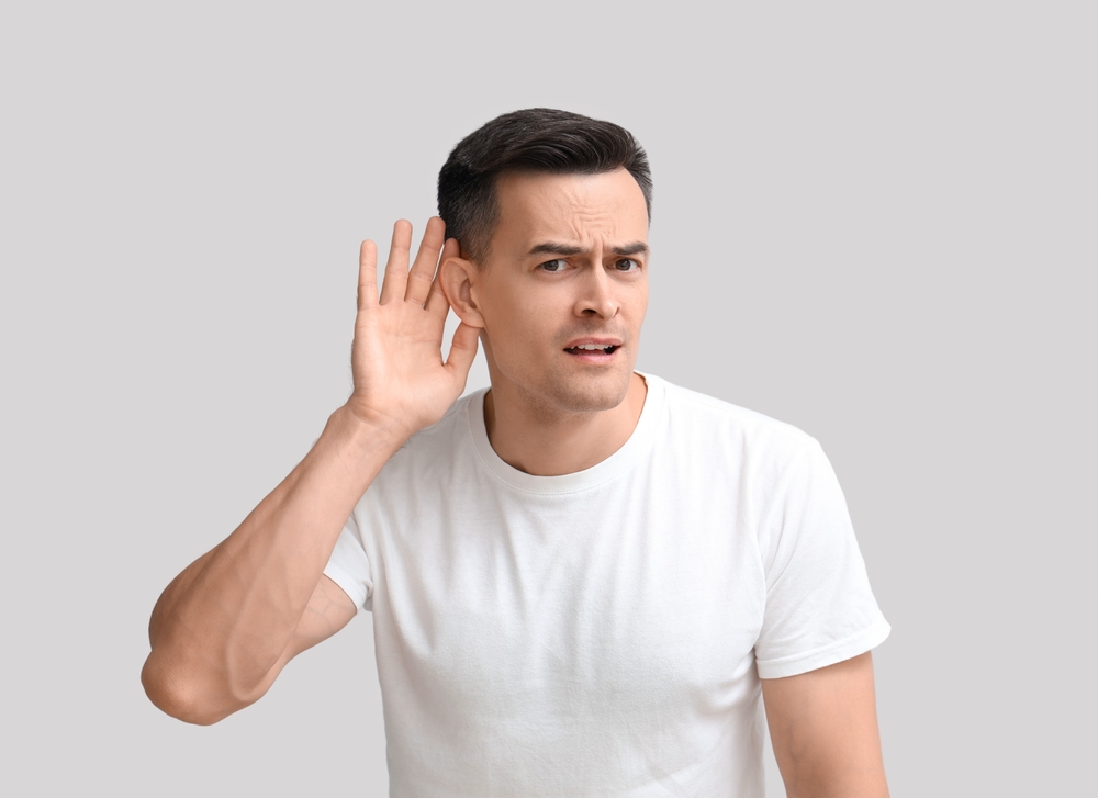 A man in a white t-shirt holds his hand to his ear and looks slightly confused or concerned, as if trying to hear something better. The background is plain light gray.