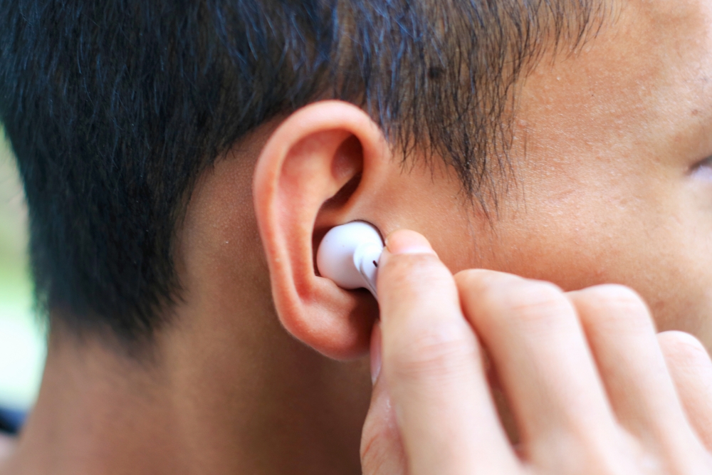 A close-up of a person inserting a white wireless earbud into their ear, with their hand gently holding the earbud. The person has short dark hair and tan skin.