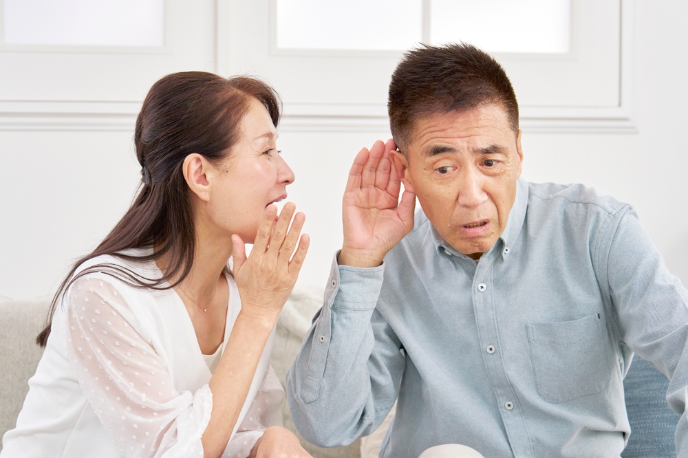 A woman is leaning close to a man, speaking softly into his ear. The man is cupping his ear and leaning in, appearing to have trouble hearing her. Both are sitting indoors and engaged in conversation.