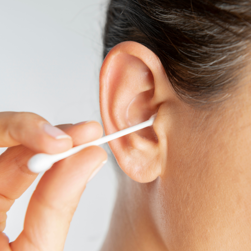 A person cleaning their ear with a cotton swab. The close-up image shows the side of their face, focusing on the ear and hand holding the swab.