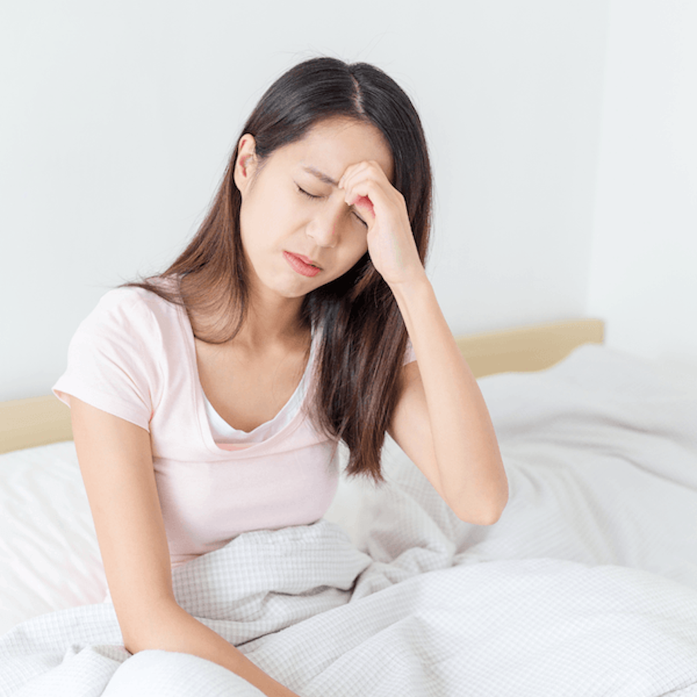 A woman sits on a bed with her eyes closed, holding her forehead and appearing to be in pain or discomfort, possibly with a headache.