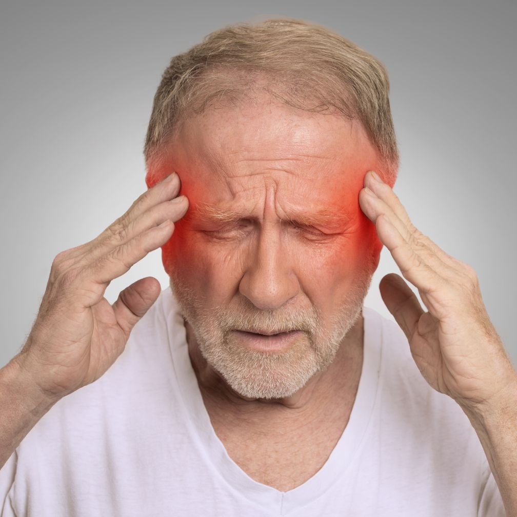 Older man with gray hair and beard holding his temples, eyes closed, and appearing to be in pain. Red highlights on both sides of his head indicate a headache or migraine. He wears a white shirt, with a gray background.