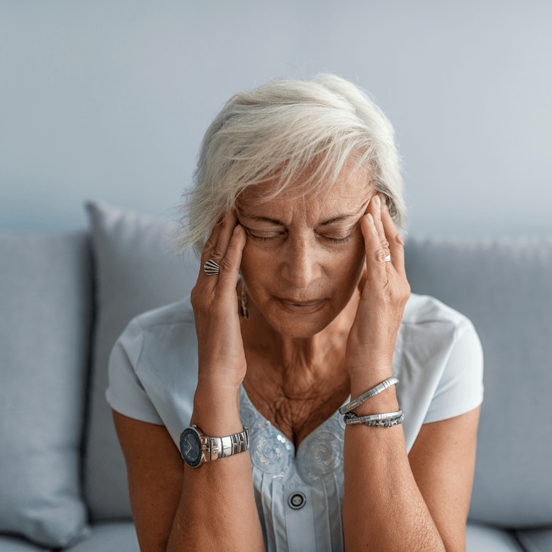 Older woman with short gray hair sits on a couch, eyes closed, pressing her hands to her temples, appearing to have a headache or be in distress.