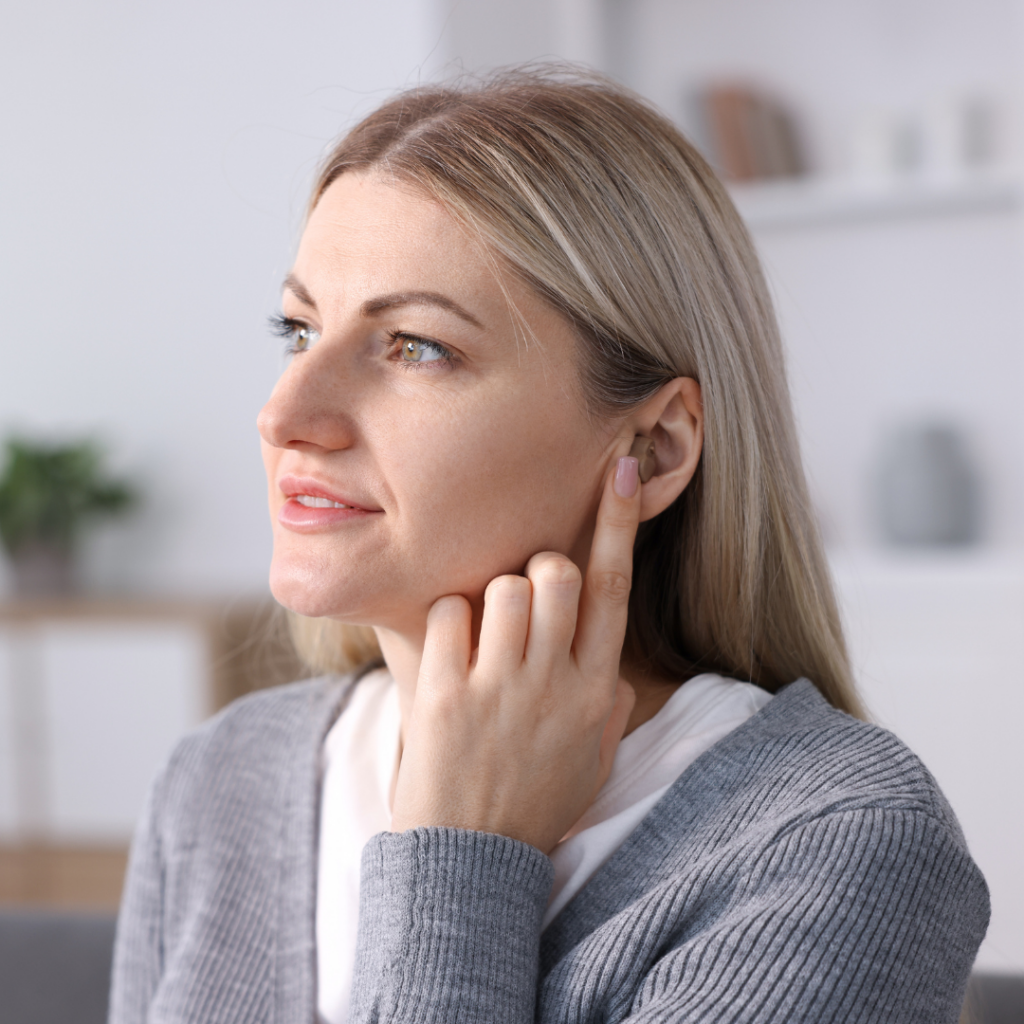 A woman with blonde hair touches her right ear with her finger, appearing thoughtful. She is indoors, wearing a gray cardigan, with a blurred background showing shelves and plants.