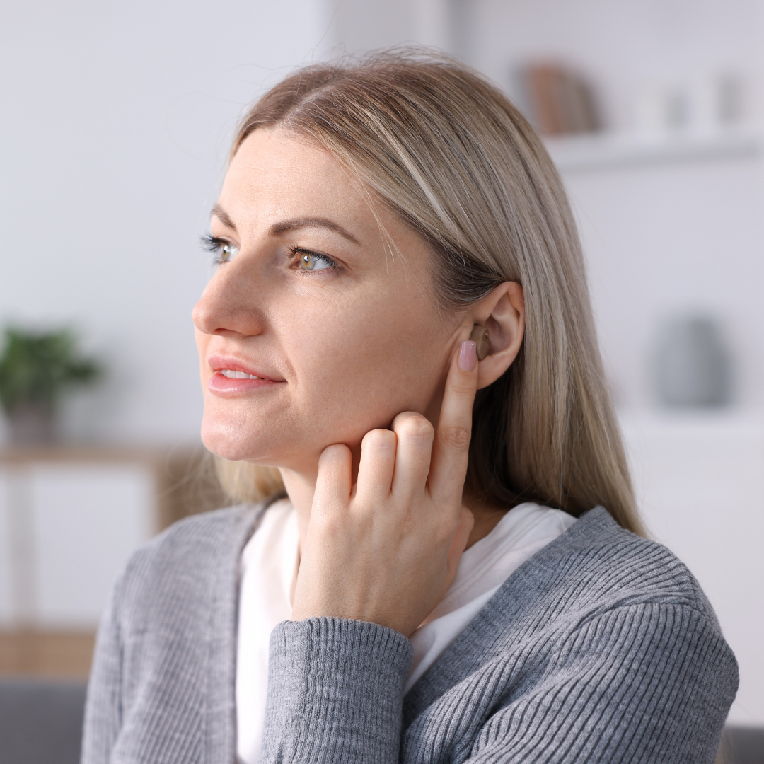A woman with blonde hair touches her right ear with her finger, appearing thoughtful. She is indoors, wearing a gray cardigan, with a blurred background showing shelves and plants.