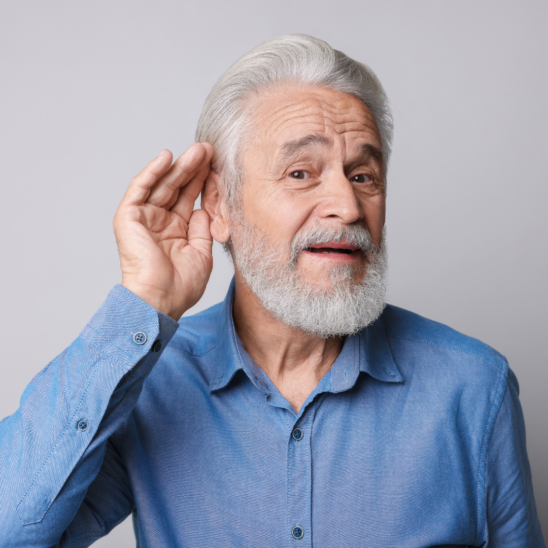 Older man with gray hair and beard, wearing a blue shirt, holds his hand to his ear and tilts his head, suggesting he is trying to hear something better, against a plain light background.